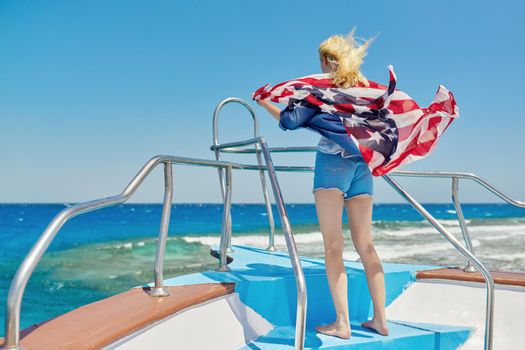 Young slender blonde woman in denim clothes with the flag of America on her shoulders, back view, in the sea on a yacht