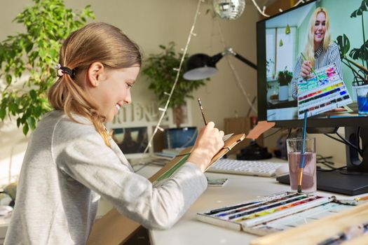 Creative online drawing lesson. Girl child drawing with watercolors, looking at computer monitor, talking with the teacher young artist