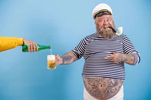 Cheerful mature sailor man with overweight and smoking pipe holds mug while woman pours tasty beer on light blue background in studio