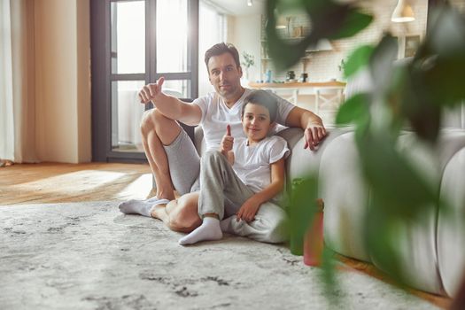 Full length portrait of smiling man sitting on floor with boy in cozy appartment and gesturing thumbs up