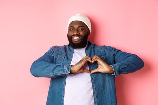 Happy african-american man showing heart sign, I love you gesture, smiling at camera, pink background.
