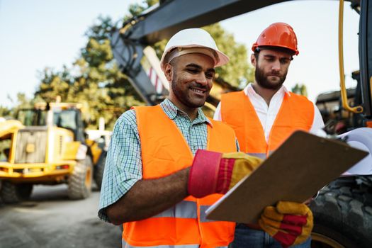 Two men engineers in workwear discussing their work standing against construction machines