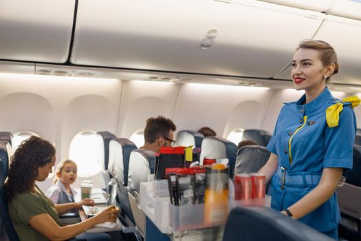 Female flight attendant serving food to passengers on aircraft. Hostess walking with trolley on aisle. Travel, service, transportation, airplane concept