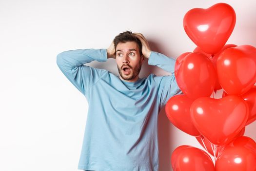 Boyfriend grab head in hands and panic over Valentines day gifts, looking sideways with alarmed face, standing near hearts balloons and thinking of present for lover, white background.