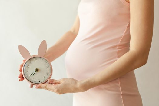 Closeup of a pregnant woman in pink dress holding bunny shaped alarm clock near her belly-maternity concept on white background