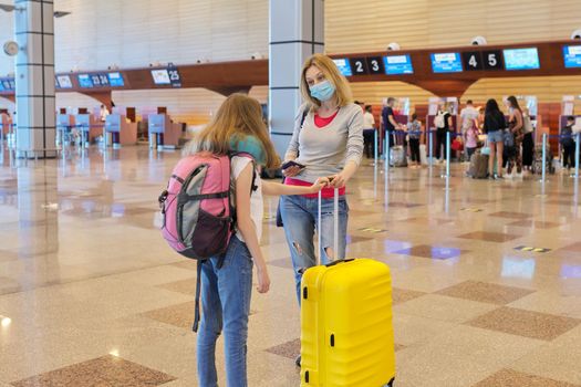 Family mother and daughter child tourists with suitcase backpacks, in protective face medical masks waiting for their plane flight at the airport terminal, check-in counter background.