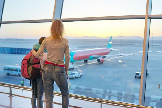 Airport passengers, standing with their backs family mother and daughter child looking at planes in panoramic window, awaiting boarding, copy space