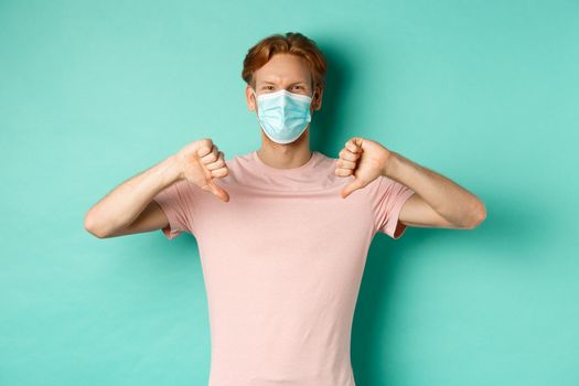 Covid-19, pandemic and lifestyle concept. Young man with red hair in face mask, showing thumbs down, dislike or disapprove something, standing over turquoise background.