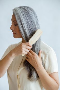 Languid middle aged Asian woman brushes natural hoary hair posing on light grey background in studio. Mature beauty lifestyle