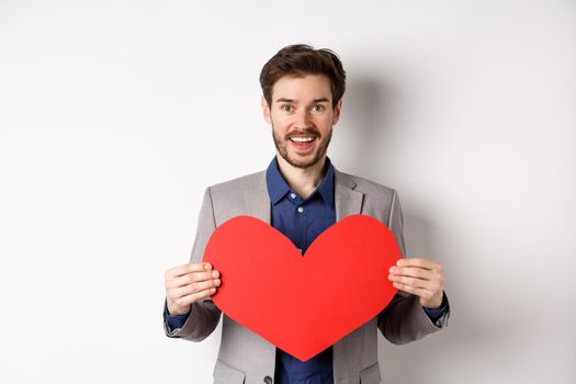 Happy man confessing in love, showing heart cutout and smiling at camera, standing in suit on romantic date with lover, white background.