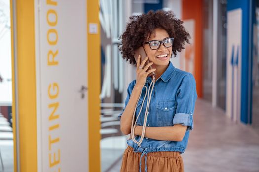 Portrait of smiling Afro American woman in glasses standing in office corridor while talking on smartphone