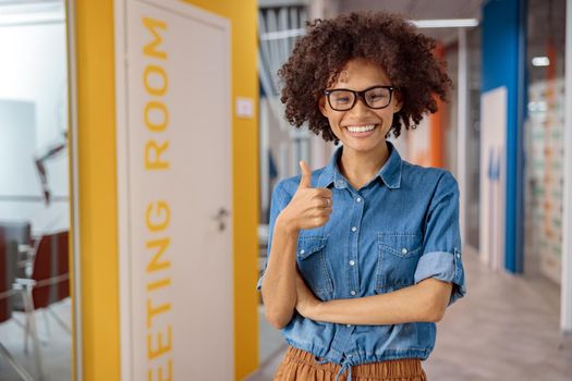 Happy Afro American woman in glasses posing in office corridor and showing thumb up