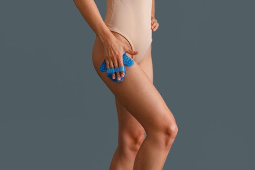 Close up shot of woman in beige bodysuit massaging her thigh with palm shaped massage glove body massager isolated over gray background. Beauty, body care concept