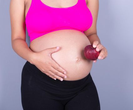 close-up belly of pregnant woman with red apple on gray wall background