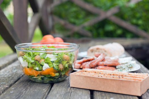 Fresh healthy salad with olives and feta chees at big bowl on wooden table. Baked cookies, bread with sausages at the picnic time. Tasty fresh food