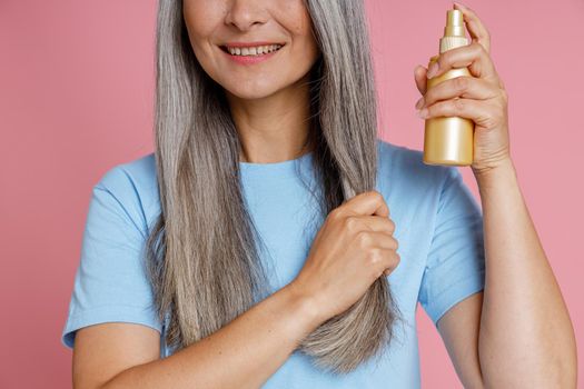 Smiling middle aged woman applies spray product onto grey hair lock standing on pink background in studio closeup. Mature beauty lifestyle