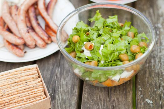 Fresh healthy salad with olives and feta chees on wooden table. Baked crisps with sausages at the picnic time. Tasty fresh food