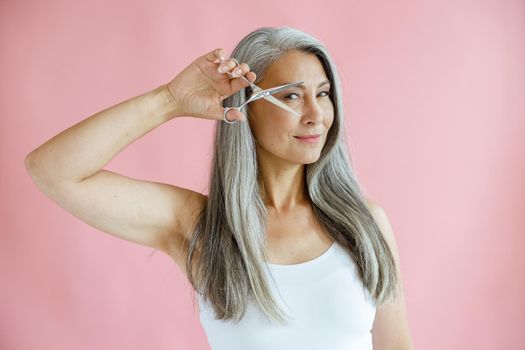 Beautiful middle aged Asian lady with long grey hair holds open scissors near face on pink background in studio. Mature beauty lifestyle