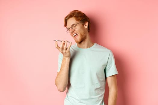 Happy young man with red hair, smiling pleased while record voice message on smartphone, talking to virtual assistant, standing over pink background.