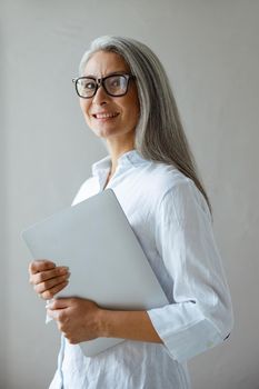 Positive long haired mature Asian lady wearing white blouse holds contemporary laptop standing near grey wall in studio