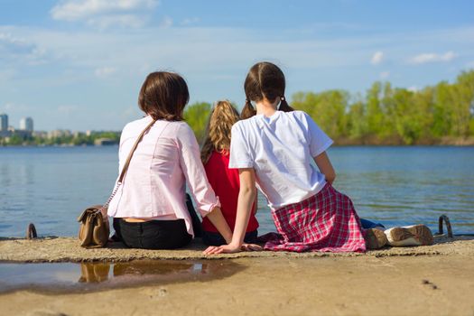 Outdoors portrait of mother and two daughters. Watching the water, back view. Background nature, park, river.