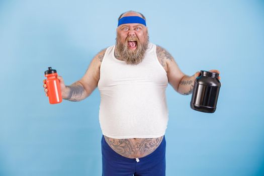 Excited middle aged plump man with headband holds bottles of drink and protein supplement posing on light blue background in studio