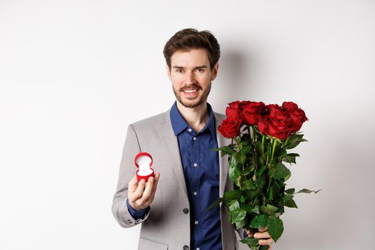 Smiling boyfriend making a marriage proposal, standing with engagement ring and bouquet of red roses, going on romantic Valentines day date, white background.