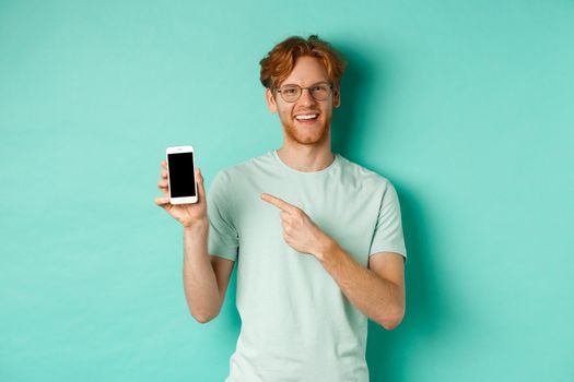 Attractive young man with red beard and hair pointing finger at blank smartphone screen, showing online promotion or app, smiling at camera, turquoise background.