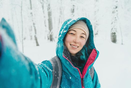 Smiling young woman doing selfie in snowy forest in winter, looking at camera