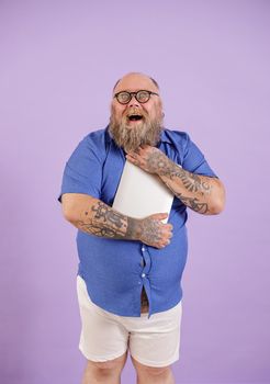 Excited bearded obese man in tight blue shirt with glasses embraces modern laptop standing on purple background in studio