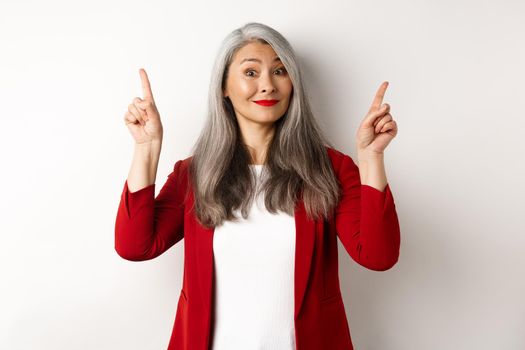 Elegant asian senior lady in red blazer and makeup, pointing fingers up and smiling, showing advertisement, white background.