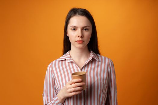 Young brunette woman looking at camera and holding paper cup against orange background, close up