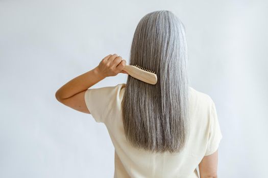 Lady in yellow blouse brushes straight silver hair standing on light grey background in studio backside view. Mature beauty lifestyle