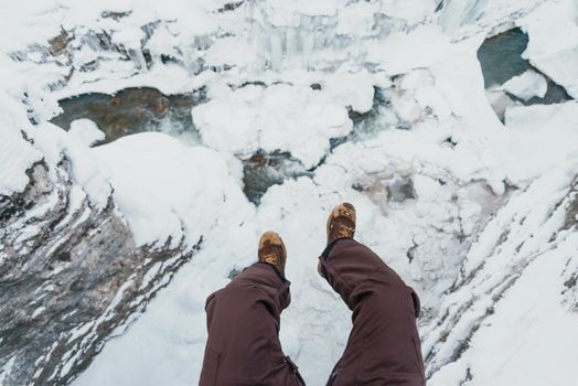 Young man sitting above the river in winter, view of legs. POV image.