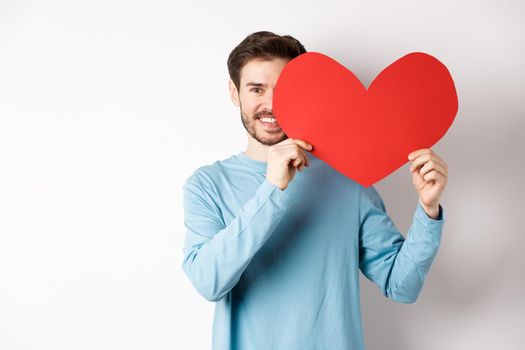 Smiling handsome man holding romantic red heart over half of face, surprise girlfriend on Valentines day, making love confession, standing over white background.