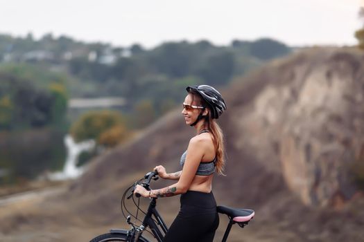 Fit young woman wearing sportswear standing with her bicycle on rocky background at autumn day