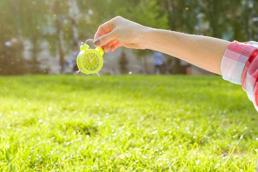 Alarm clock in female hand, background green grass in park, sunlight, copy space