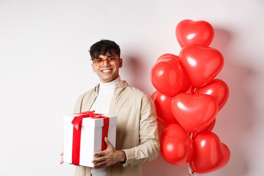 Valentines day and romance concept. Man in love prepare surprise gift for lover, holding present in box and standing near red hearts gesture, white background.
