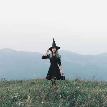 Beautiful young woman in costume of witch walking on autumn meadow. Halloween theme