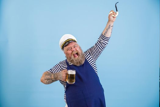 Bearded obese man in sailor suit with apron holds glass mug of beer and raises up smoking pipe on light blue background in studio