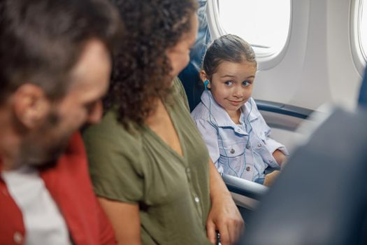 Cute little girl wearing earphones while sitting on the plane, listening to music, looking at her parents, traveling together with family. Travel, vacation concept