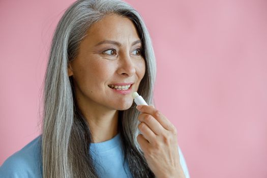 Positive silver haired Asian woman in blue t-shirt applies lip balm on pink background in studio, space for text. Mature beauty lifestyle