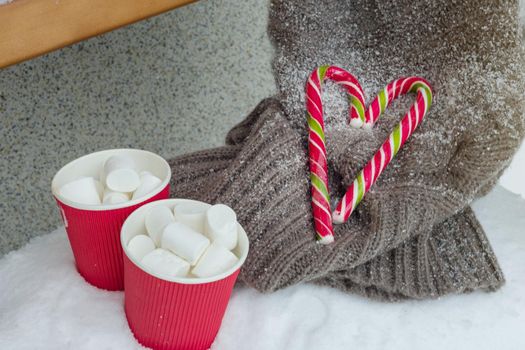 Winter background - two cups of coffee with marshmallow, two christmas candy canes - heart, knitted sweater on the snow bench