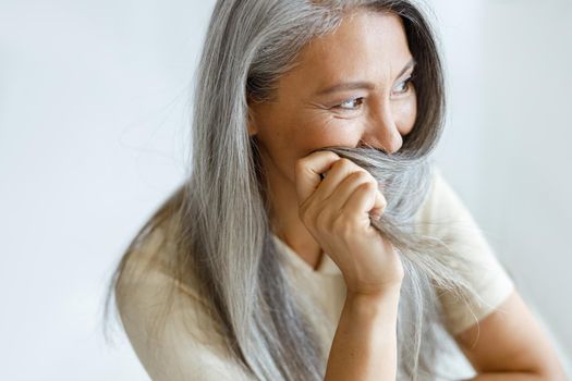 Playful middle aged Asian woman hides face with beautiful grey hair sitting on light background in studio. Mature beauty lifestyle