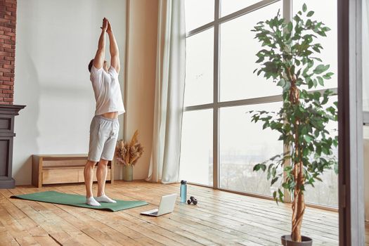 Male is practicing asana near window in living room while watching workout on notebook during quarantine