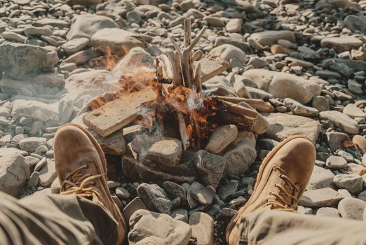 Young man sitting near the bonfire on pebble coast, view of legs, pov