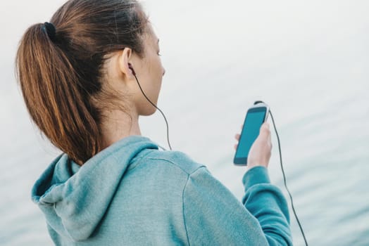 Fitness young woman in headphones listening music on her smartphone for workout on background of sea