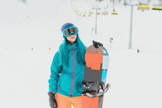 Young woman with blue hair wearing in protective sunglasses standing with snowboard on ski resort, looking at camera