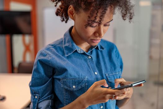 Concentrated young woman typing message while using phone and looking on screen in the office