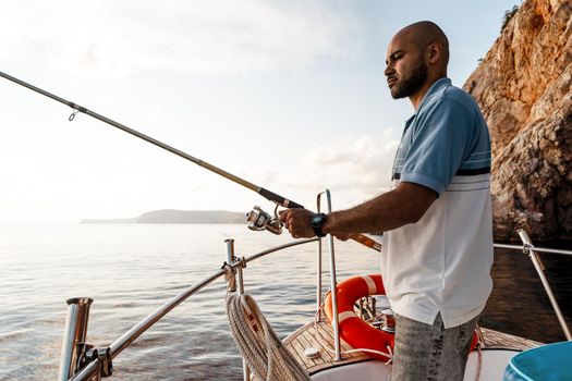 Young african american man standing with fishing rod on a sailboat fishing in open sea on sunset, close up